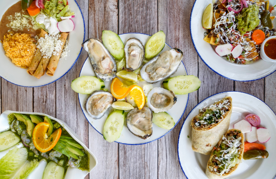A vibrant spread of Mexican and seafood dishes on a rustic wooden table, featuring oysters on the half shell, tacos, burritos, nachos, and a fresh salad with cucumber slices and orange garnish.