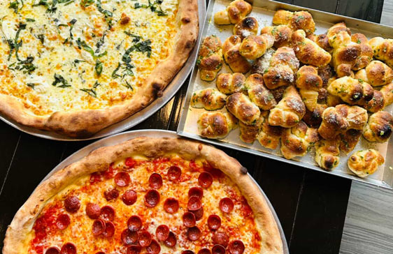 Top‑down spread of four large pizzas—white spinach, pepperoni cup, and plain red—framing a tray piled high with golden garlic knots.