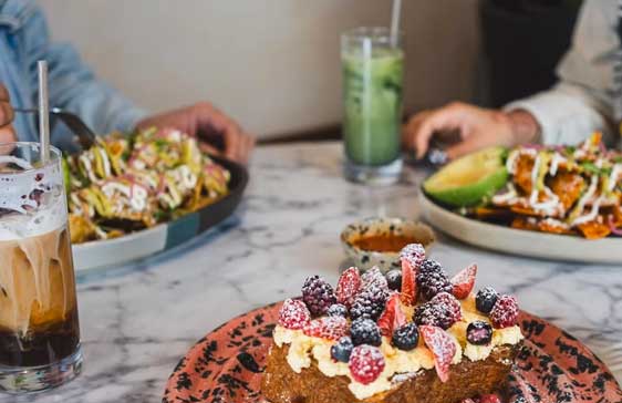 Brunch spread on a marble table at Amor A Mi featuring a berry-topped French toast with whipped cream, a plate of nachos drizzled with sauce, and a dish with avocado and crispy tortilla. Two iced drinks, including an iced coffee and a matcha latte, accompany the meal while two people enjoy their food.
