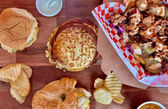 A top-down view of a wooden table filled with various sandwiches, crispy potato chips, a creamy dipping sauce, and a loaded fries dish in a checkered paper tray. The spread includes croissant and brioche-style buns.