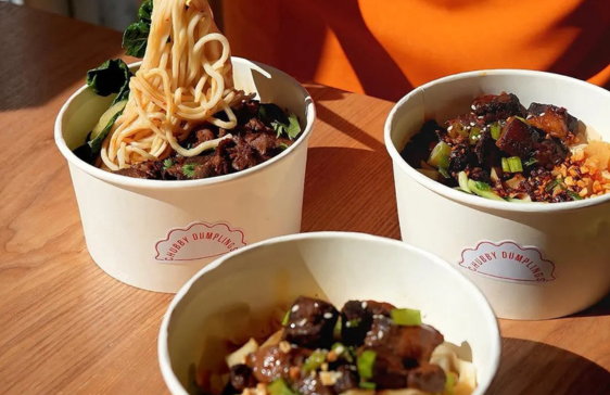 Three bowls of noodles topped with braised meat, vegetables, and scallions on a wooden table.