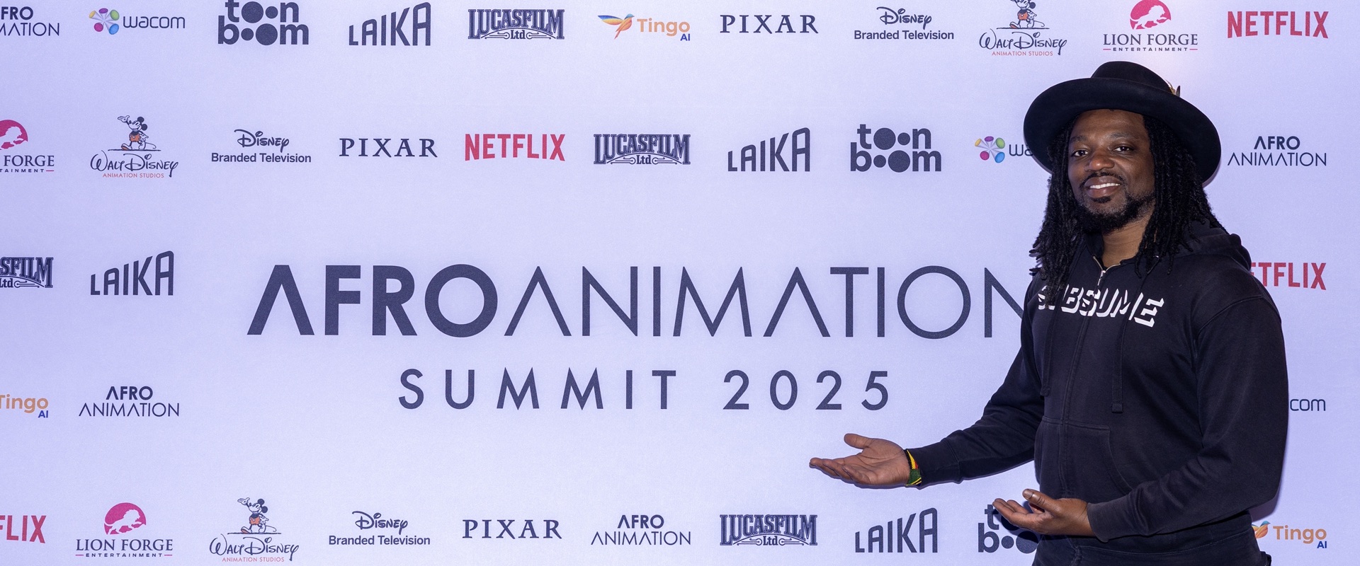 A guest in a black hat gestures toward the AfroAnimation Summit 2025 logo on a sponsor wall of animation studio logos