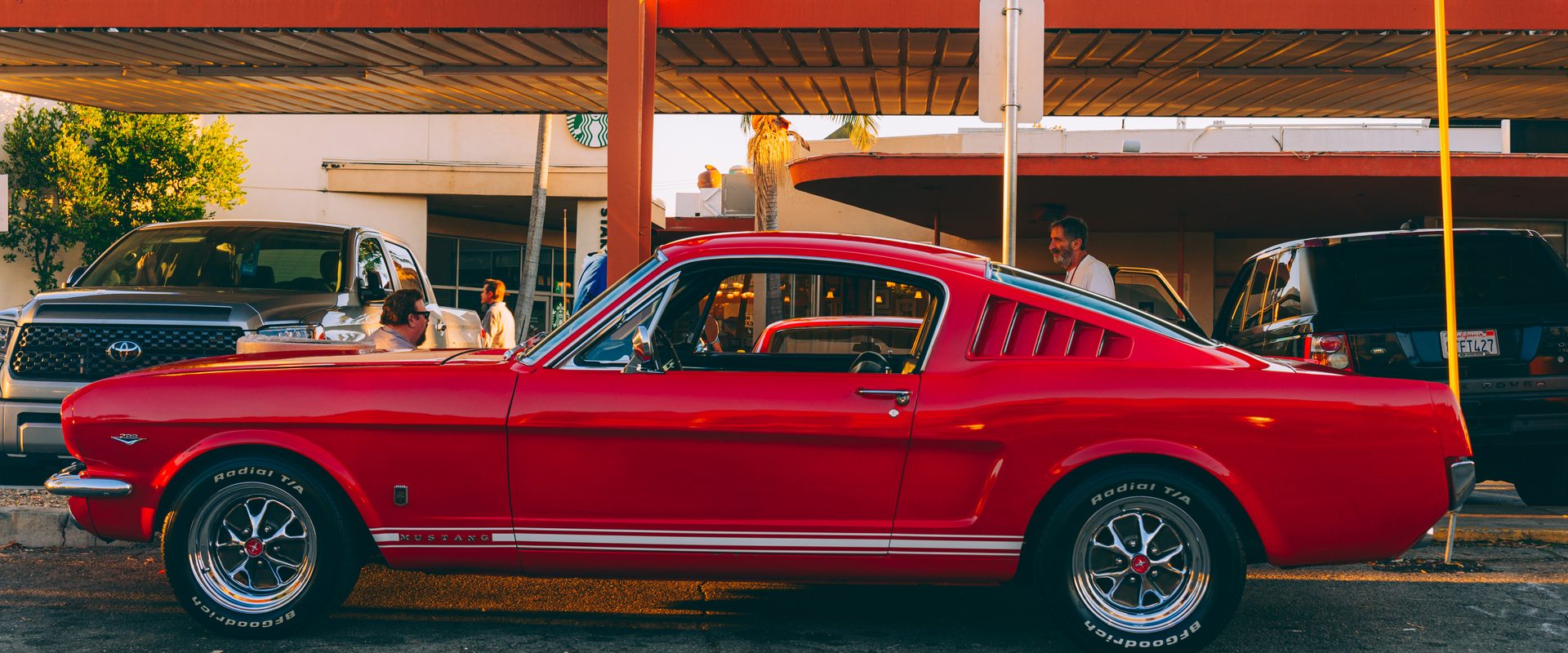 A red 1960s Ford Mustang fastback parked under the Bob's Big Boy drive-in canopy at golden hour