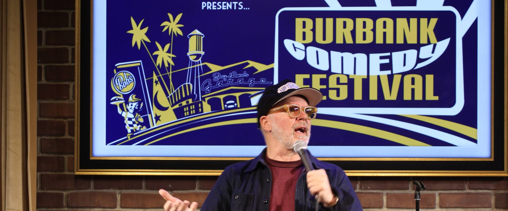 A comedian in a cap and glasses performs under the big Burbank Comedy Festival screen at Flappers Comedy Club