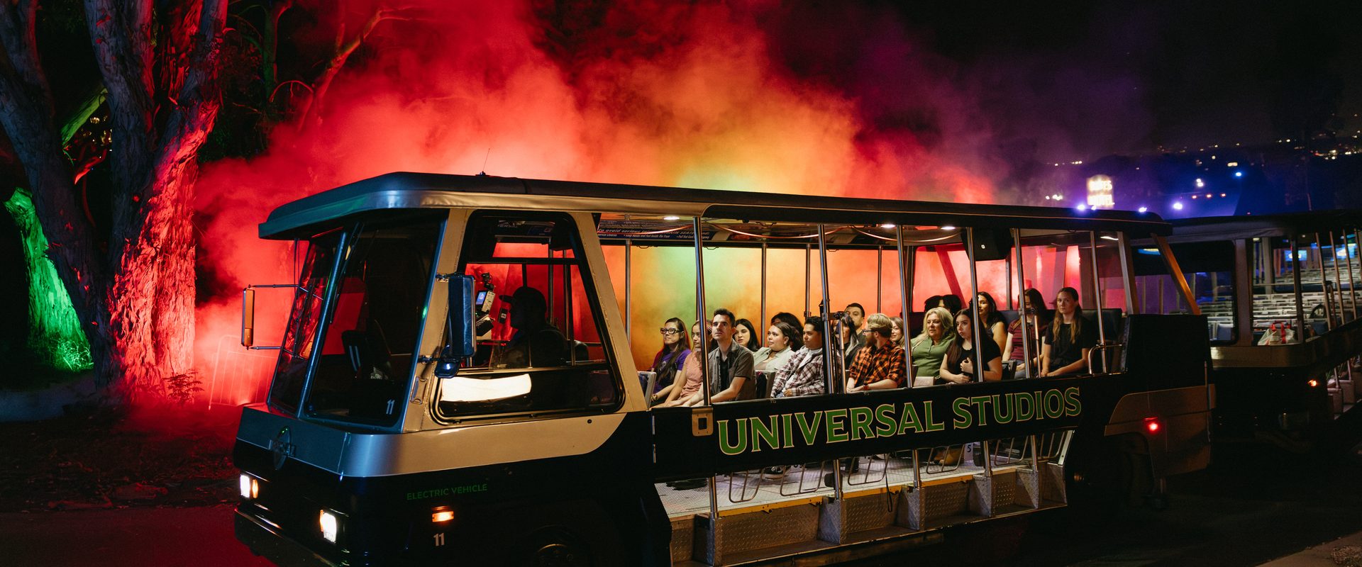 Universal Studios tram full of riders rolls past red smoke and uplit trees on the Terror Tram at Halloween Horror Nights