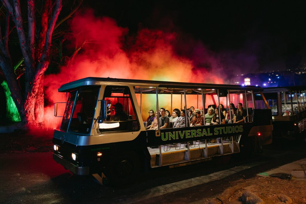 Universal Studios tram carries riders past red smoke and uplit trees on the Terror Tram at Halloween Horror Nights