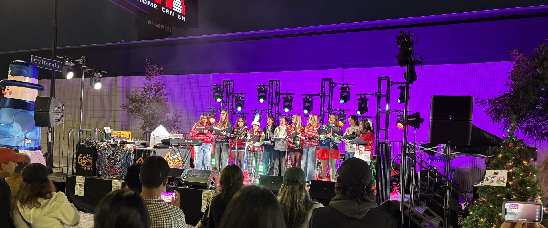 A choir in holiday sweaters sings on a purple-lit stage on Magnolia Boulevard as the crowd films on their phones