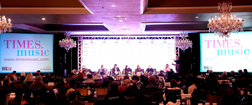 A MUSEXPO panel on stage in a chandelier-lit ballroom, with Times Music logos on the big screens either side