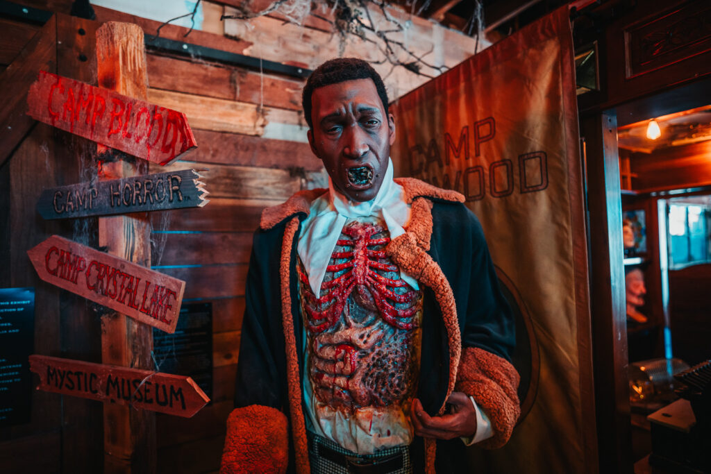 Camp Horror signpost pointing to Camp Blood, Camp Crystal Lake and the Mystic Museum beside a ghoulish figure in Burbank