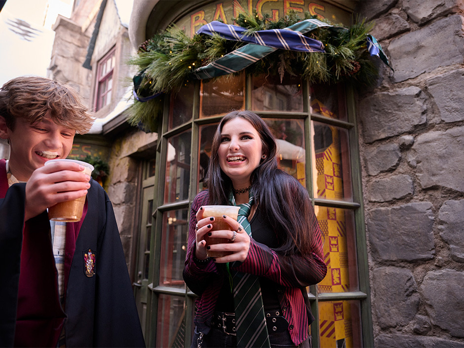 Two visitors in Hogwarts robes holding mugs of Butterbeer in Hogsmeade