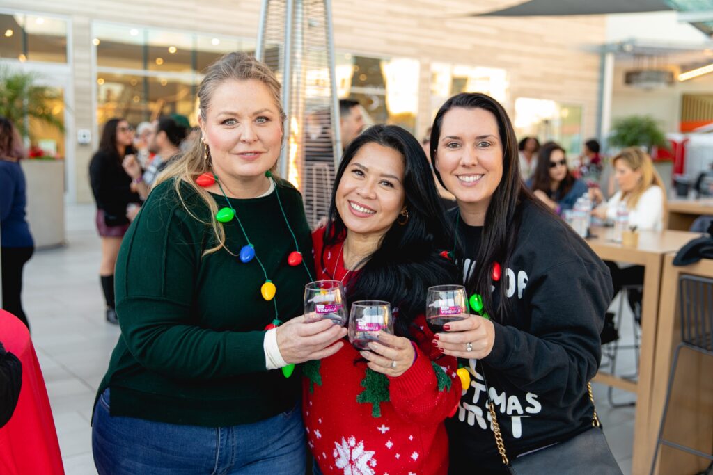 Three friends in holiday sweaters holding Wine Walk tasting glasses