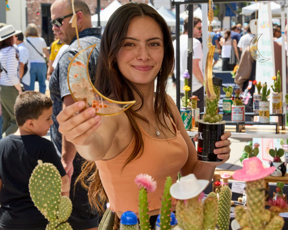 A vendor holds up handmade jewelry beside cactus planters at her booth